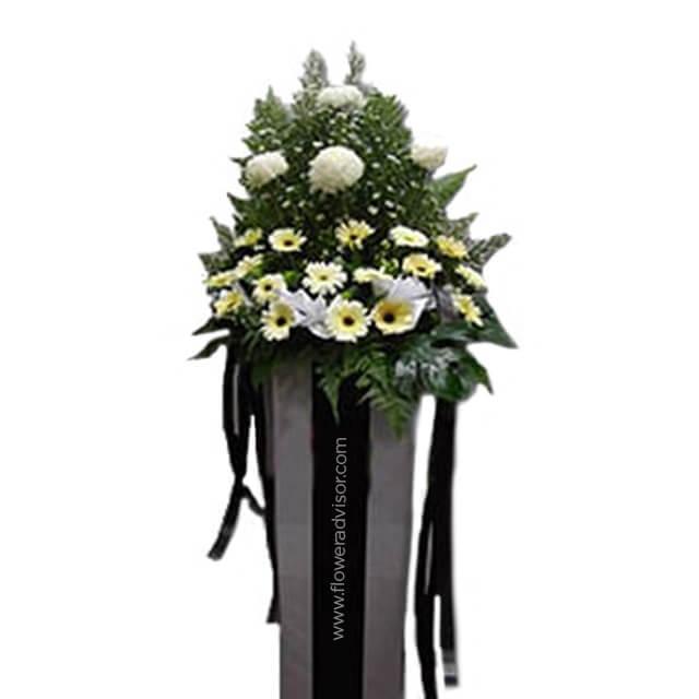 Standing flower arrangement with white and yellow chrysanthemums and daisies, accented by greenery, displayed in a tall box with black ribbons.