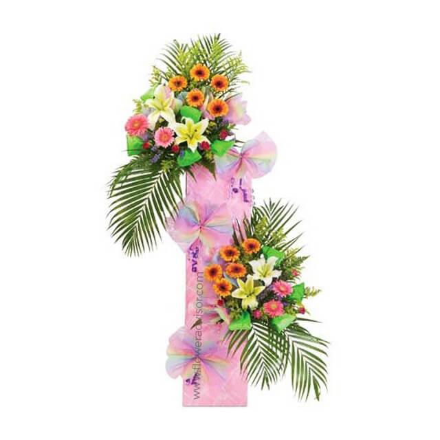 Tall floral arrangement with pink lilies, orange gerbera daisies, green palm leaves, and pink ribbon bow on white background