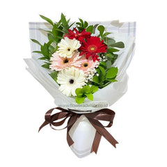 Bouquet of mixed gerbera daisies in red, white, and pale pink with green foliage wrapped in white paper and tied with a brown ribbon