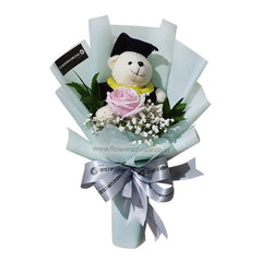 Graduation bouquet with a teddy bear in black cap and gown, pink rose, baby's breath, green leaves, and silver ribbon