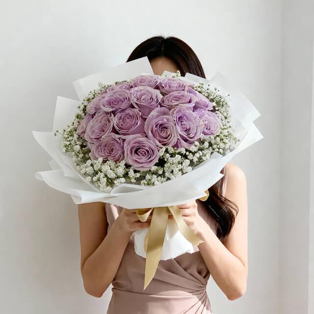Woman holding large bouquet of fresh lavender roses and white baby's breath wrapped in white paper with gold ribbon