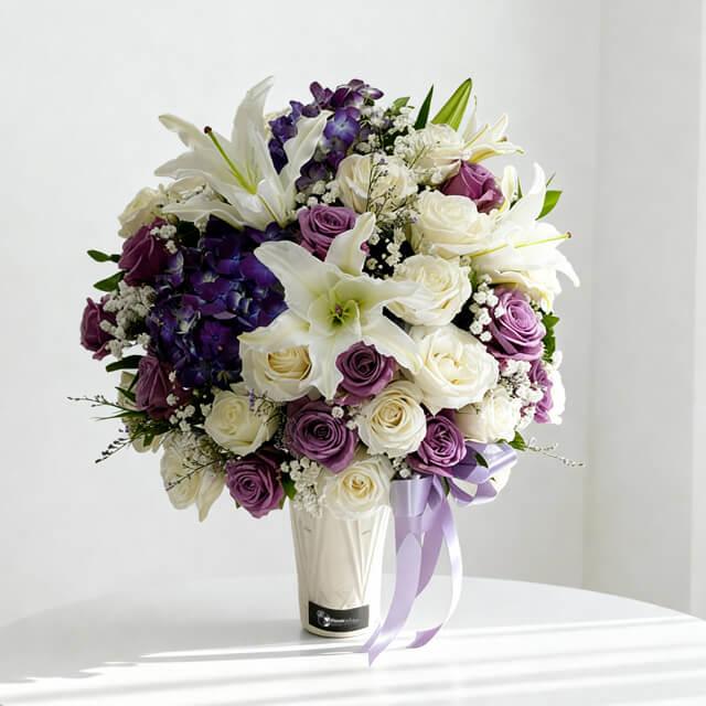 Elegant bouquet of white lilies, purple and white roses, and purple hydrangeas in a white vase with lavender ribbon