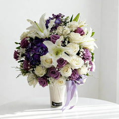 Elegant bouquet of white lilies, purple and white roses, and purple hydrangeas in a white vase with lavender ribbon