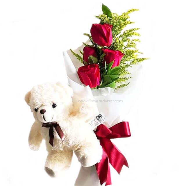 Bouquet of three red roses with green foliage wrapped in white paper, tied with a red ribbon, accompanied by a small white teddy bear with a brown ribbon bow