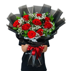 Hand holding a bouquet of red roses and white baby's breath wrapped in black paper with red ribbon