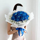 Person holding a bouquet of blue roses wrapped in white paper with babyβs breath, tied with a blue ribbon