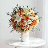 Elegant bouquet of orange and cream roses with white lilies and greenery in a white vase on a round table