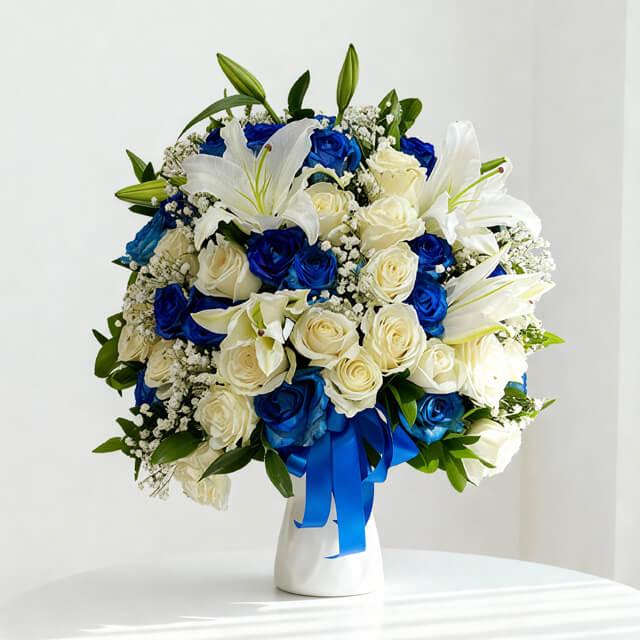 Elegant bouquet of white lilies and cream roses with vibrant blue roses and greenery in white vase
