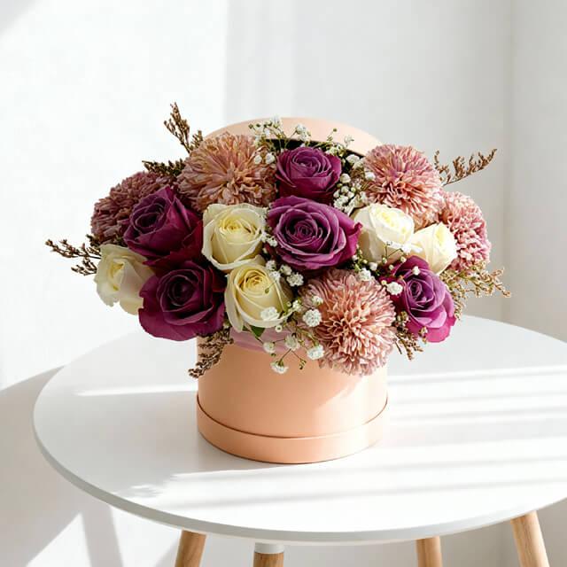 Elegant bouquet of purple and cream roses with pink chrysanthemums in a peach round box on white table
