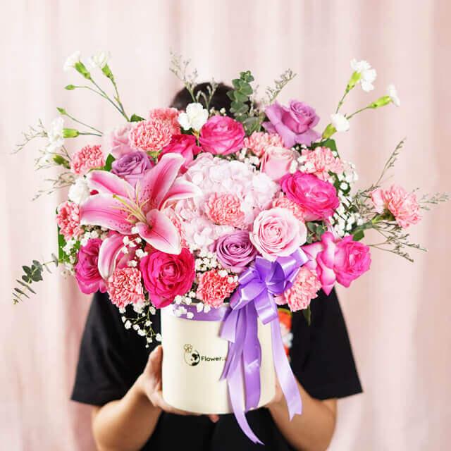 Bright pink and purple mixed flower bouquet with lilies, roses, carnations, and greenery in white vase with purple ribbon