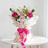 Elegant bouquet of pink and red roses with white flowers and greenery, wrapped in white paper with a pink ribbon on a white table