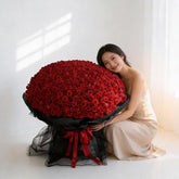 Woman in a light dress sitting beside a giant bouquet of red roses wrapped in black and red ribbon