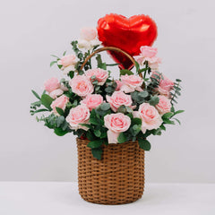 Wicker basket arrangement with fresh pink roses, green foliage, and a red heart-shaped balloon