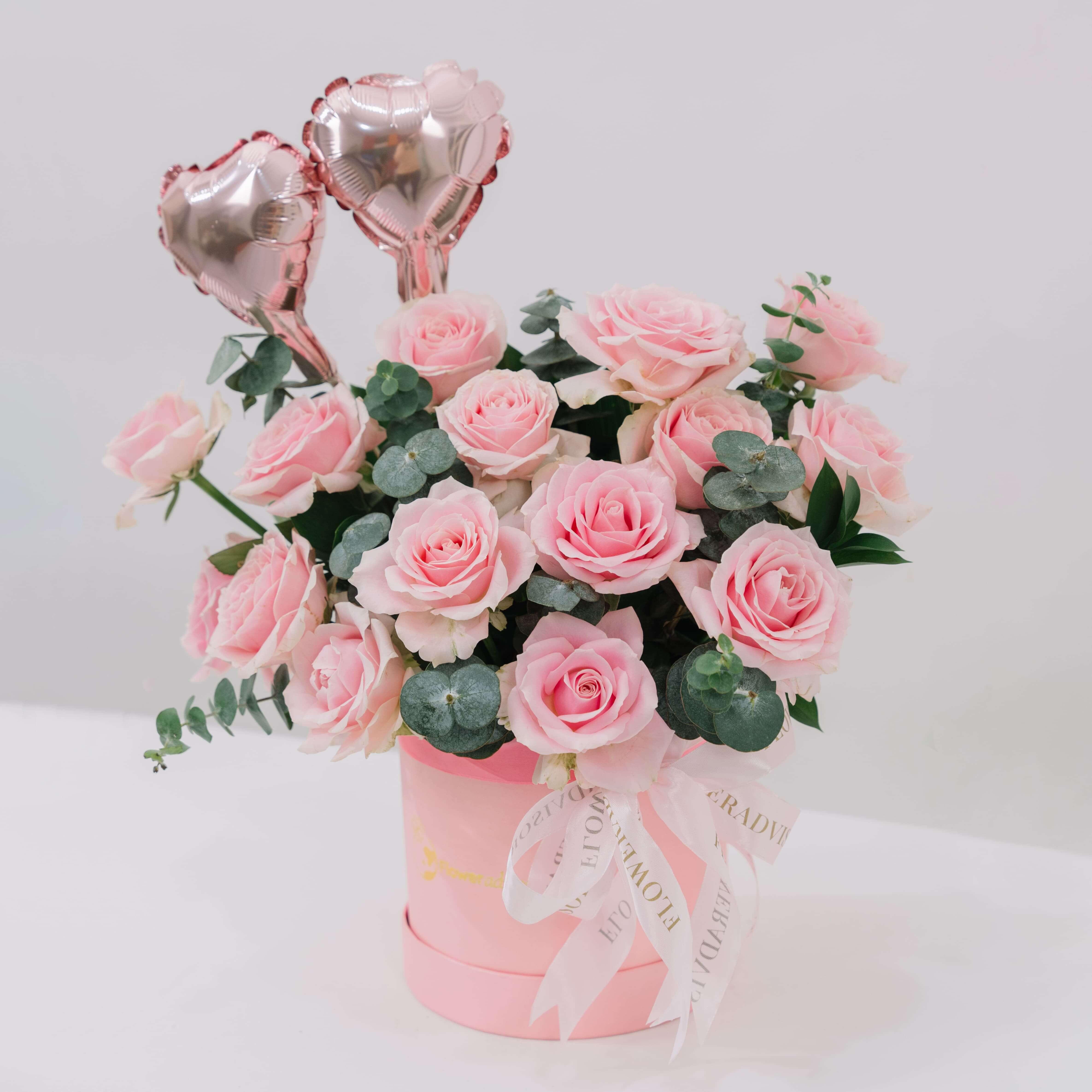 Pink roses bouquet with eucalyptus leaves in a pink box adorned with ribbon and two heart-shaped balloons