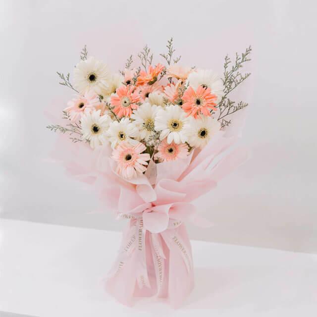 Delicate bouquet of white and peach gerbera daisies with greenery wrapped in soft pink paper tied with ribbon