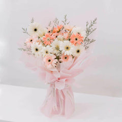 Delicate bouquet of white and peach gerbera daisies with greenery wrapped in soft pink paper tied with ribbon