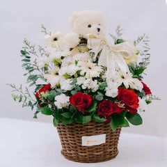 Flower basket arrangement with red roses, white flowers, greenery, and a cream teddy bear with branded ribbon