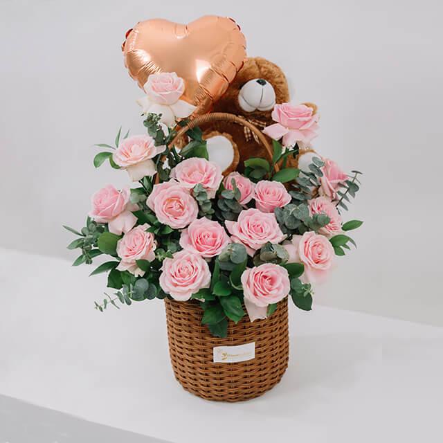 Basket of fresh pink roses with eucalyptus, a plush teddy bear, and a gold heart-shaped balloon
