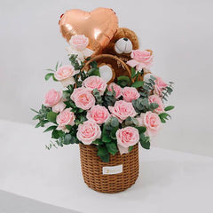 Basket of fresh pink roses with eucalyptus, a plush teddy bear, and a gold heart-shaped balloon