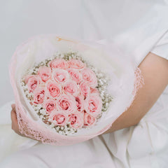 Person in white holding bouquet of light pink roses wrapped in white and pink mesh with baby's breath flowers