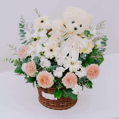 Wicker basket with pastel peach and white flower bouquet featuring eucalyptus and a cream teddy bear with FlowerAdvisor ribbon