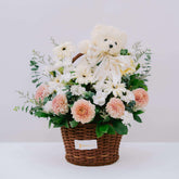 Wicker basket flower arrangement with white daisies, pink dahlias, green foliage, and a small teddy bear with FlowerAdvisor ribbon
