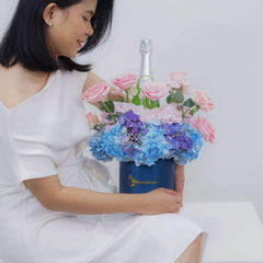 Woman in white dress holding a navy blue flower box with pink roses and blue hydrangeas, featuring a bottle in the center