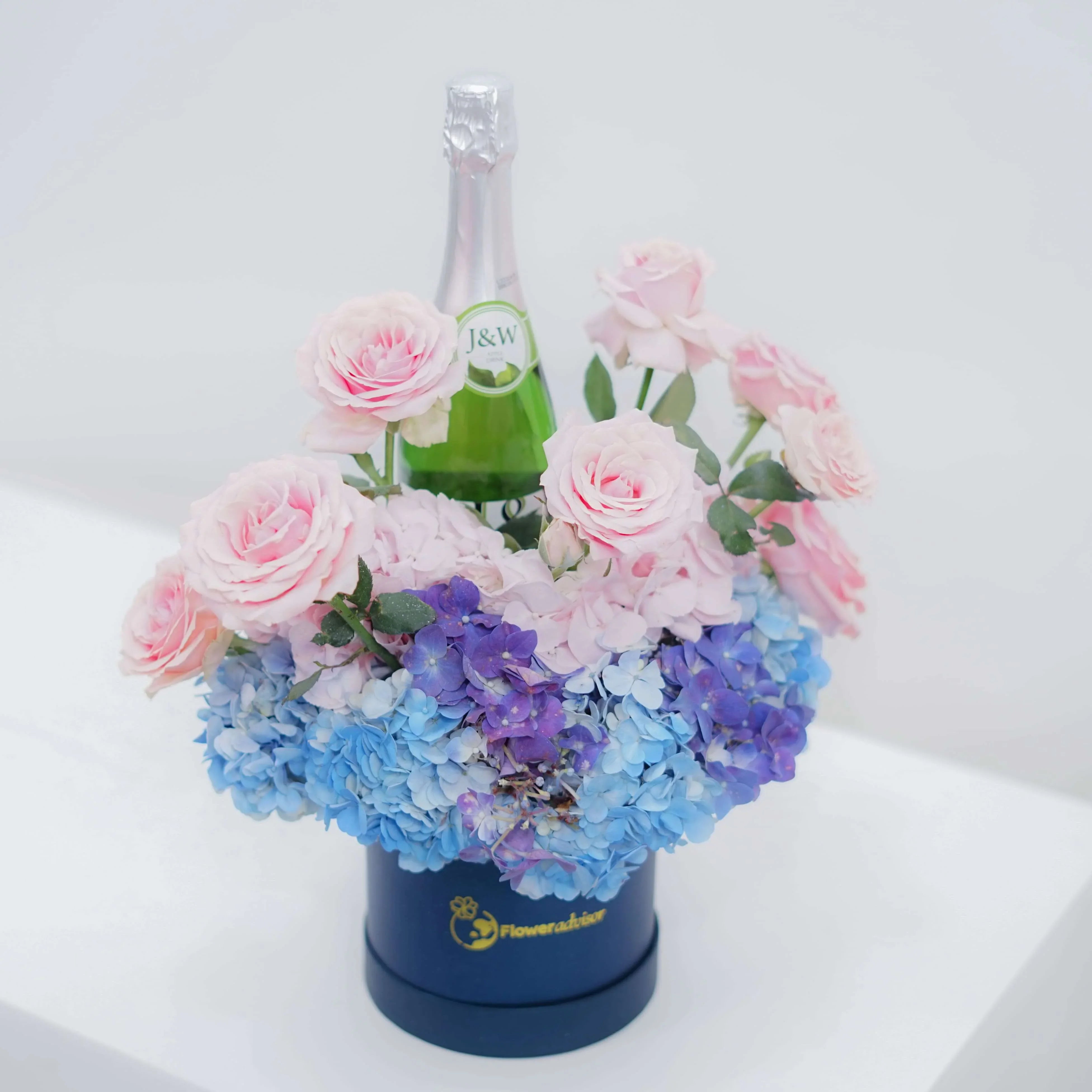 FlowerAdvisor floral arrangement with pink roses, blue hydrangeas, and a champagne bottle in a navy box