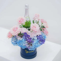 FlowerAdvisor floral arrangement with pink roses, blue hydrangeas, and a champagne bottle in a navy box