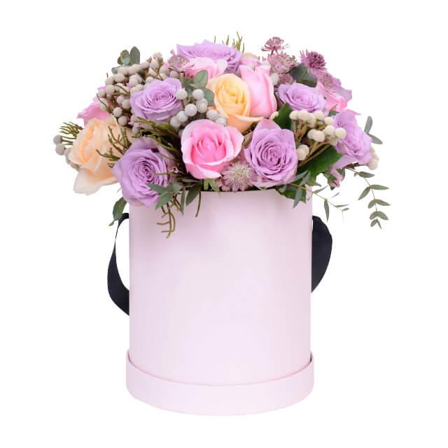 Pastel bouquet of pink, lavender, and peach roses with greenery in a pink round flower box