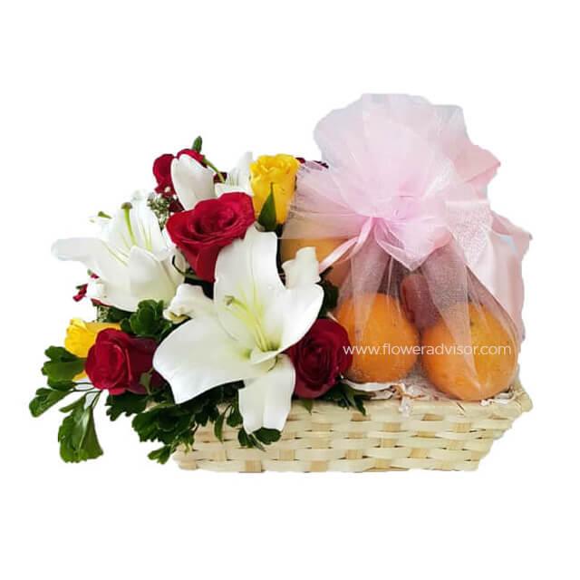 Flower basket with white lilies, red and yellow roses, and a pink netted bag containing oranges
