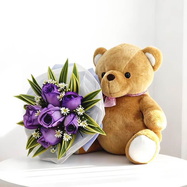 Brown teddy bear with pink ribbon holding purple rose bouquet with greenery on white surface