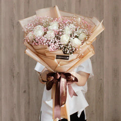Bouquet of white roses and pink baby's breath wrapped in beige paper with brown ribbon, held by person