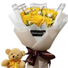 Bouquet of fresh yellow roses wrapped in white and beige paper with brown ribbon, accompanied by a small plush teddy bear with a yellow bow