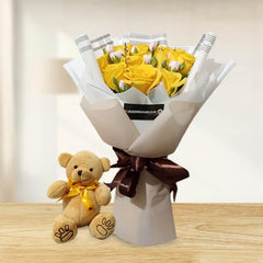 Bouquet of yellow roses and small white flowers wrapped in white paper with brown ribbon beside a small teddy bear with a yellow bow on wooden table
