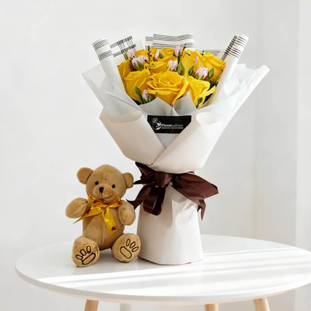Bouquet of yellow roses with pink buds wrapped in white paper and brown ribbon beside small teddy bear on round white table