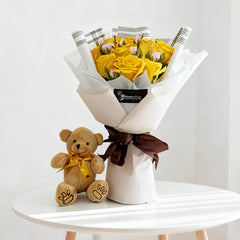 Bouquet of yellow roses with pink buds wrapped in white paper and brown ribbon beside small teddy bear on round white table