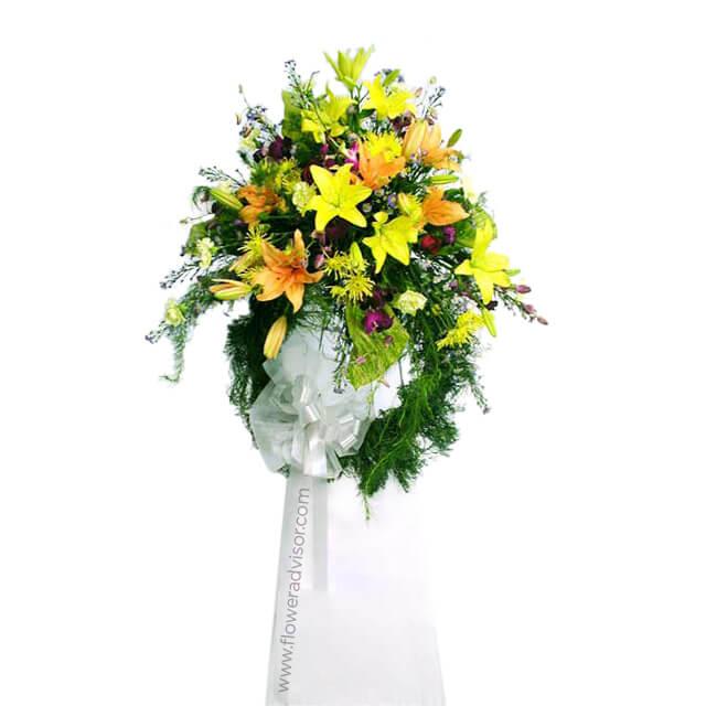 Elegant standing floral arrangement with yellow lilies, orange flowers, green foliage, and white ribbon