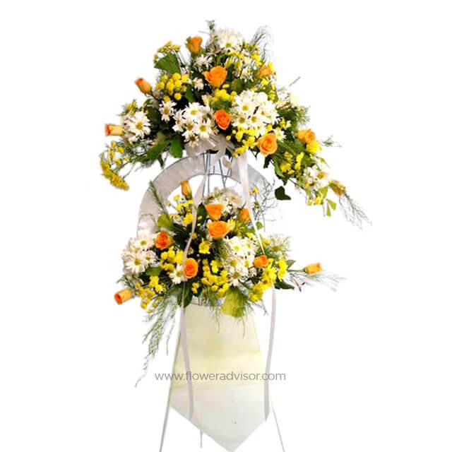 Two-tier floral arrangement with white daisies, yellow flowers, and orange roses on white stand