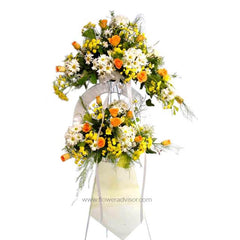 Two-tier floral arrangement with white daisies, yellow flowers, and orange roses on white stand