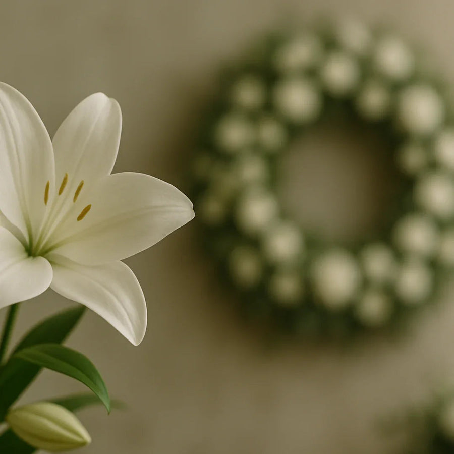 Close-up of a white lily flower with green leaves and a blurred floral wreath background