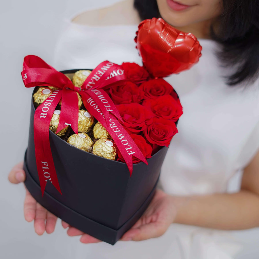 Woman holding a black heart-shaped gift box with red roses, chocolates, red ribbon, and heart balloon