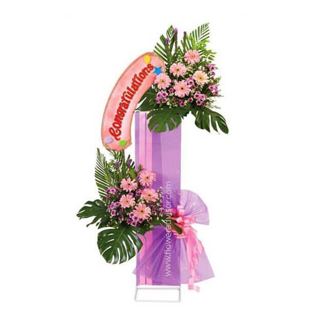 Two-tier congratulatory flower stand with pink and purple mixed flowers and a 'Congratulations' balloon.