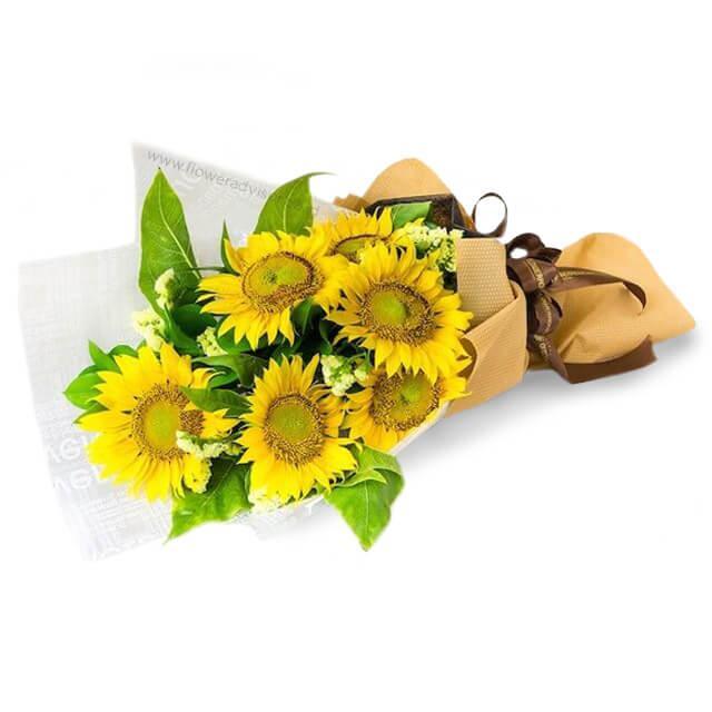 Bouquet of six fresh sunflowers with green leaves and white filler flowers wrapped in brown paper.