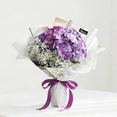 Bouquet of purple hydrangeas and white baby's breath wrapped in white paper with purple ribbon