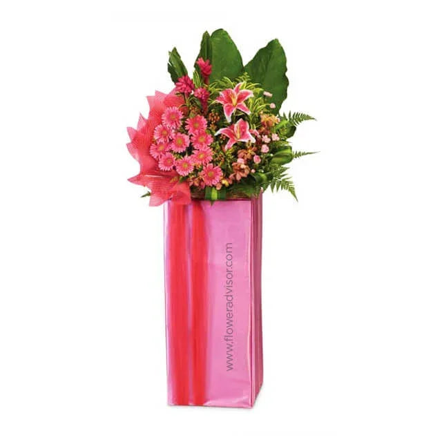 Arrangement of pink gerbera daisies and pink lilies in a tall pink bouquet box with greenery