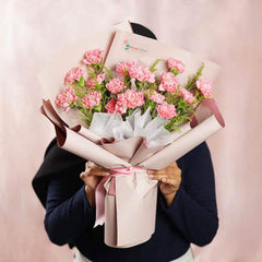 Person holding a bouquet of pink carnations wrapped in beige and brown paper with floral greenery from FlowerAdvisor Malaysia