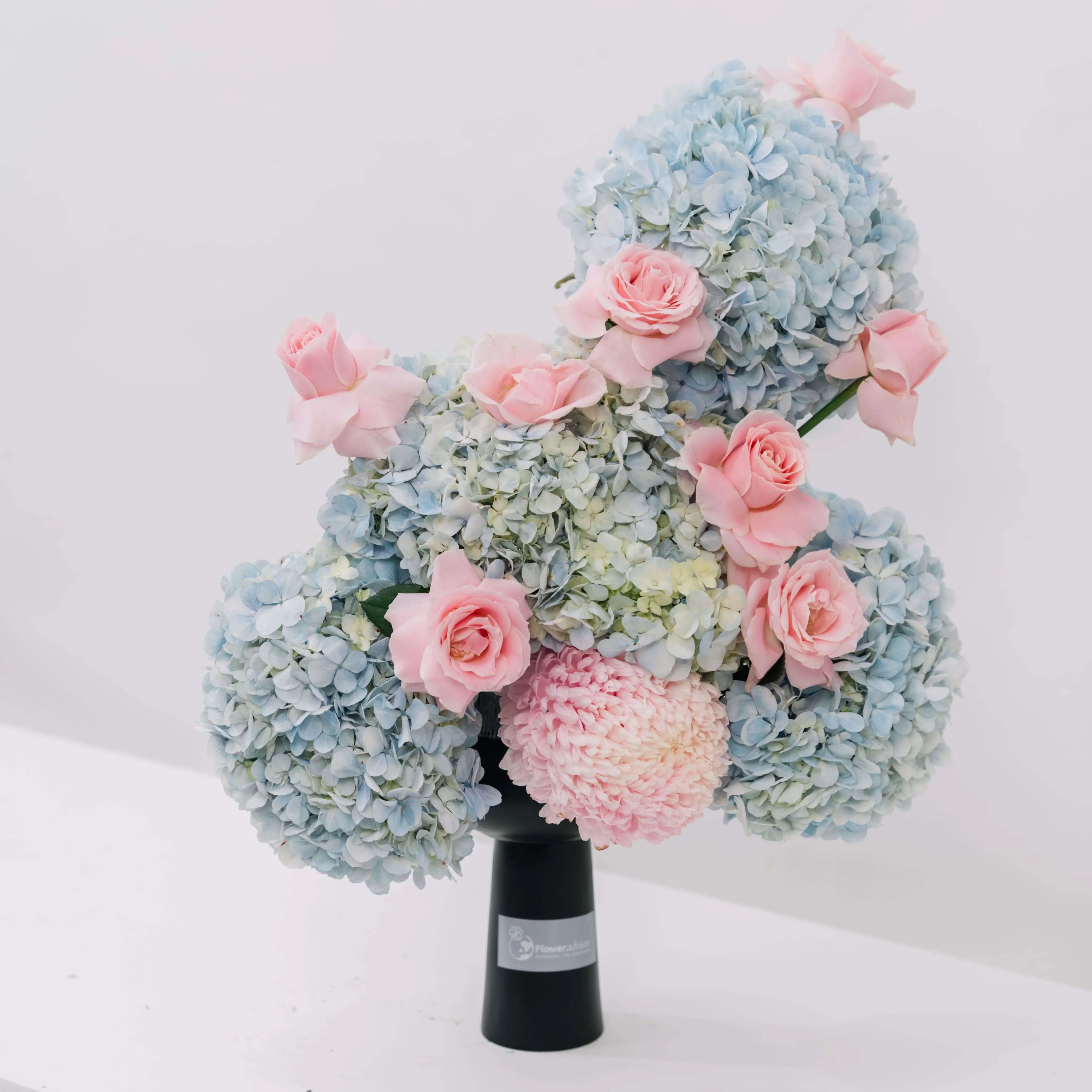 Elegant bouquet of blue hydrangeas and soft pink roses arranged in black vase on white background